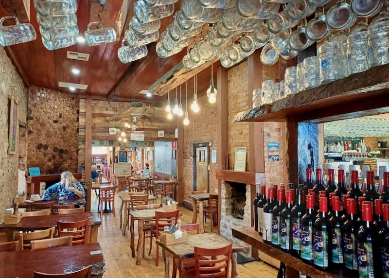 A woman sits in a restaurant with glass beer mugs overhead and wine bottles on a shelf.