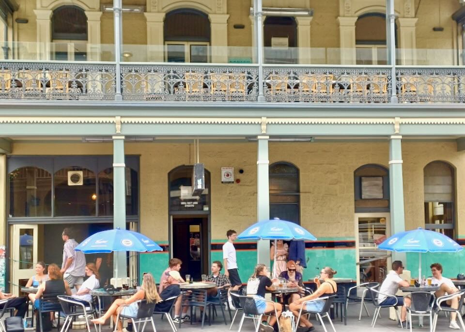 People sit outside a historic building at tables under blue umbrella and eat and drink.