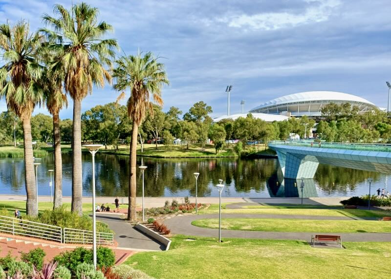 Palm trees stand over a river next to a pedestrian bridge to a white round building.