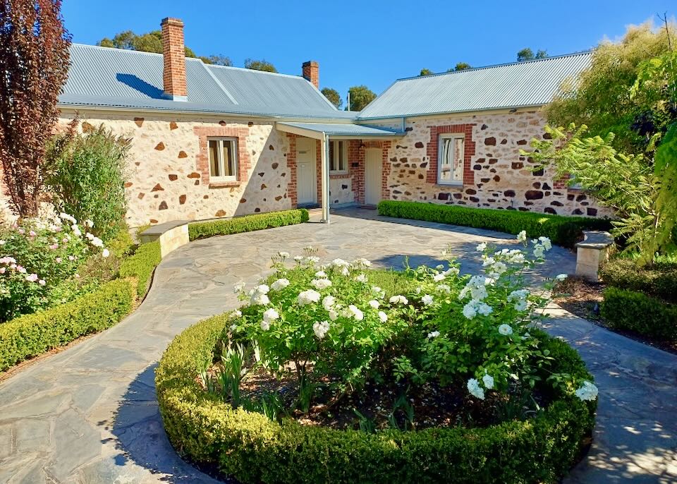 Green bushes and white flowers sit next to stone pathways to white and brown stone cottages.