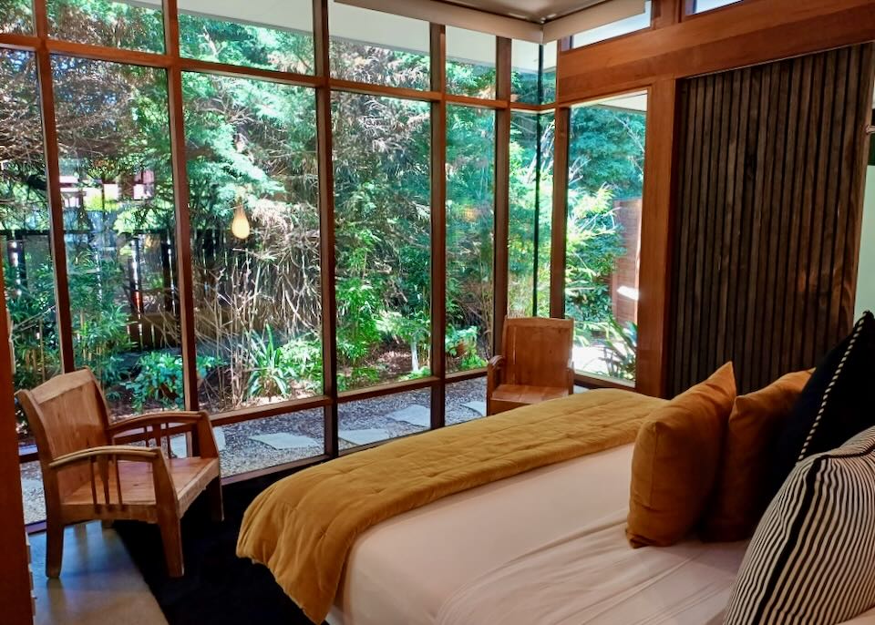 A hotel bed faces a wall of windows looking onto green trees.