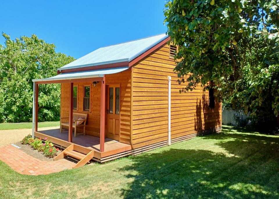 A small orange wood cabin sits on a green grass lawn with a bench and flower bed out front.