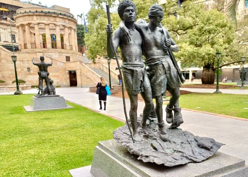 In a park a sculpture of two men, one carrying a rifle with a bandage over his eyes, leans on another man caring a walking stick.