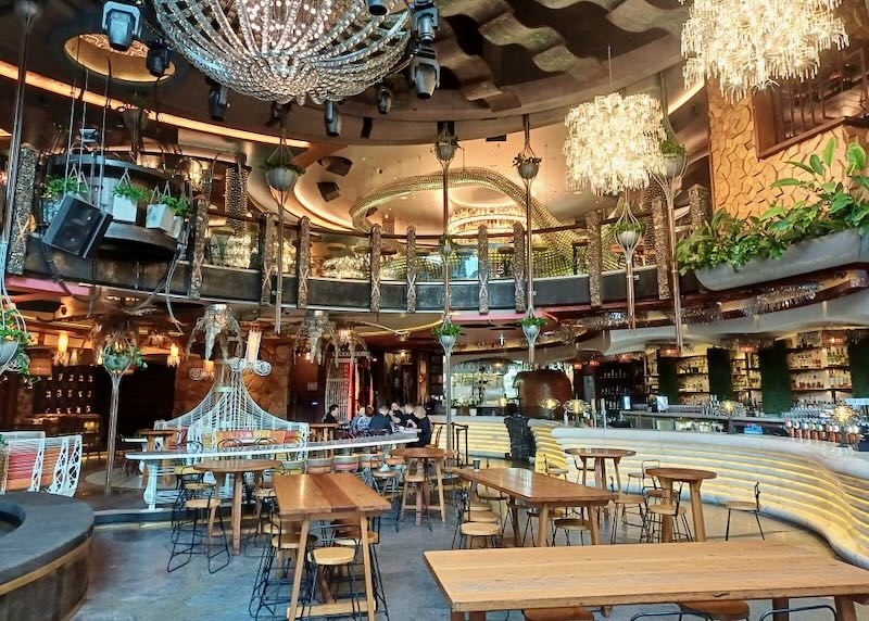 A restaurant dining room filled with multiple hanging crystal chandeliers, live plants, a curved marble bar with lights, and wood tables.