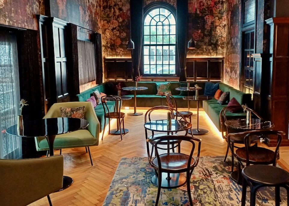 A moody bar lounge with dark green velvet benches and wood chairs in a historic wood filled room.