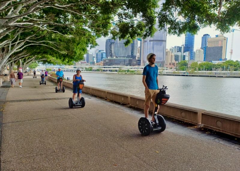 People ride Segways under trees by a river.