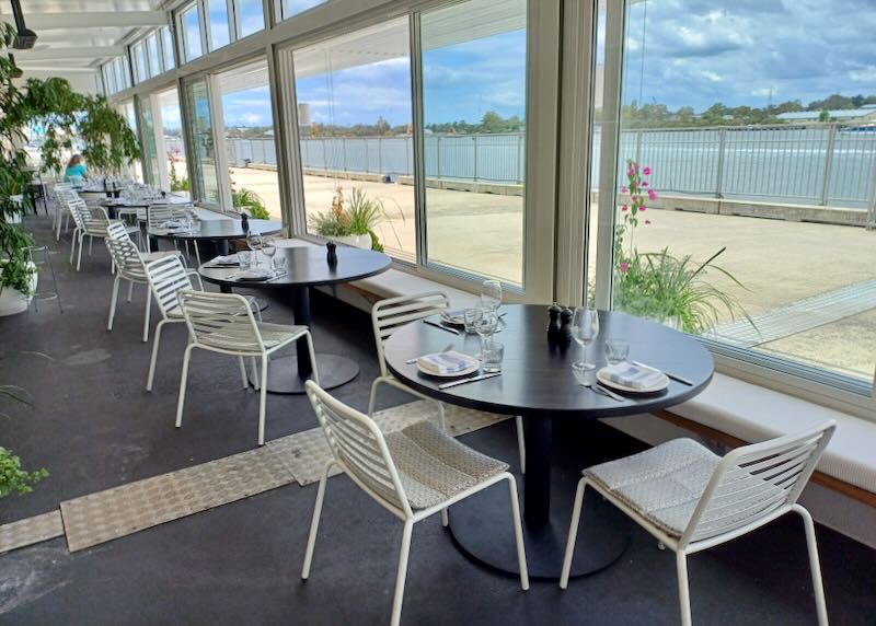 Table and chairs sit in a restaurant next to windows and views of the water.