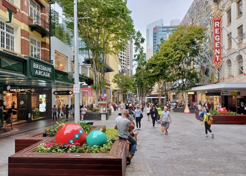 People shop in a pedestrian mall carrying bags and sitting on wood benches.