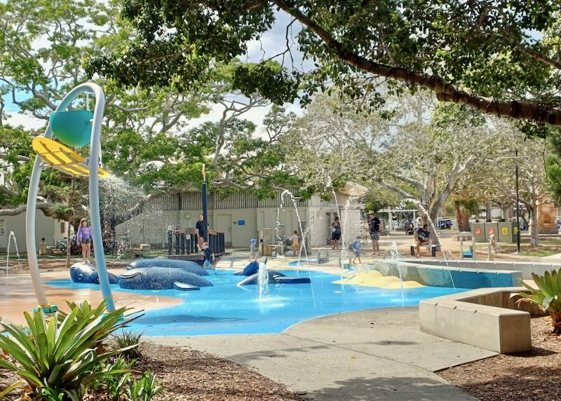 Kids play in a shallow water bed filled with sculptures of whales, fountains, and a large dumping bucket of water.