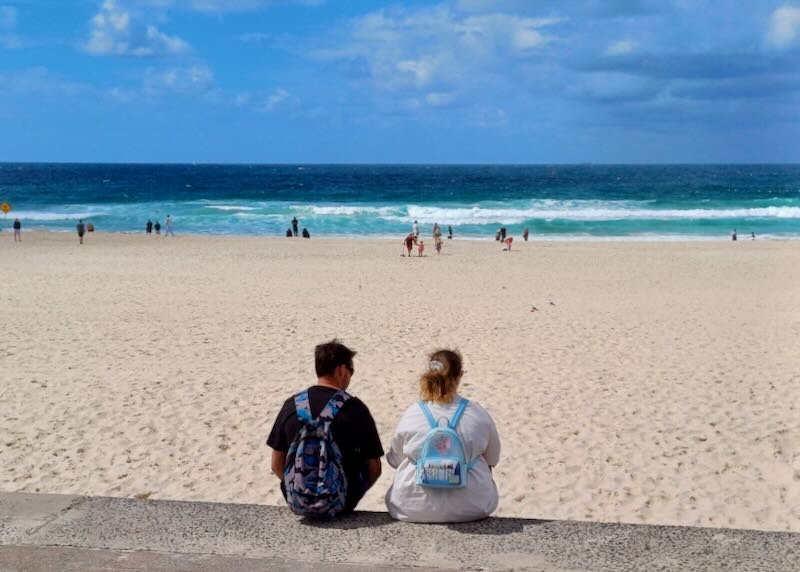 Two people sit on a retaining wall on a white-sand beach with blue ocean waves in the distance.