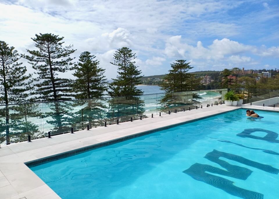 A couple hug in a roof-top pool overlooking evergreen trees and an ocean.