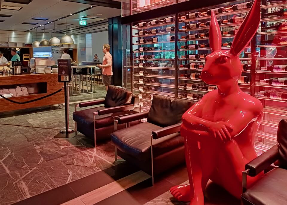 A large red sculpture of a man with a rabbit head sits between two chairs in a lobby.