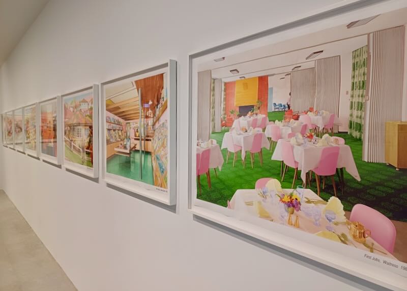 A row of pink, green, yellow, and orange paintings line a wall at an art museum.