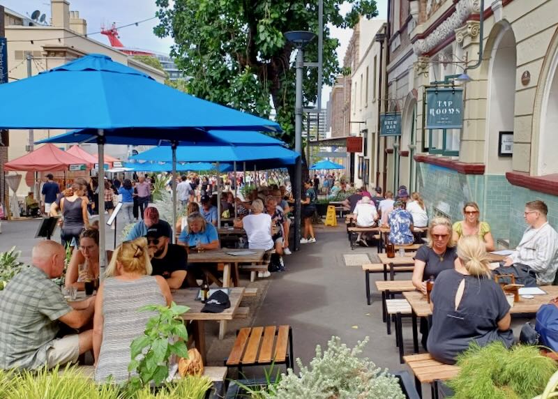 Crowds of people sit on sidewalk picnic tables and benches to watch the people go by.