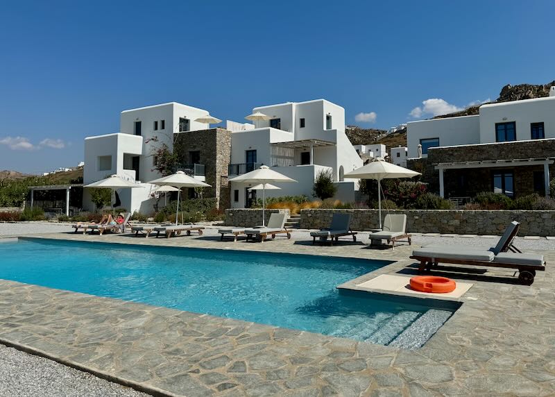 Blue swimming pool, backed by a white Cycladic-style hotel on a sunny day