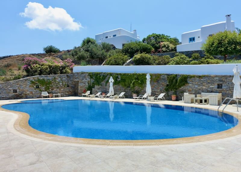 Blue swimming pool with two white hotel villas on a hill behind it