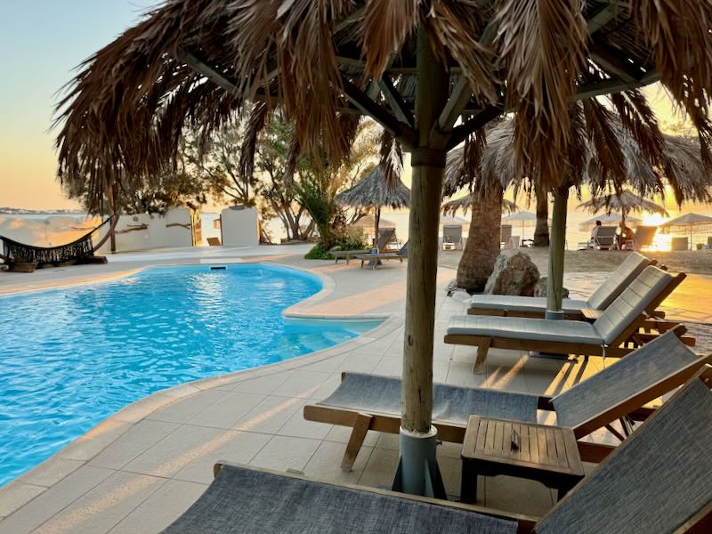 Hotel swimming pool with thatched umbrellas, set next to a sandy beach.