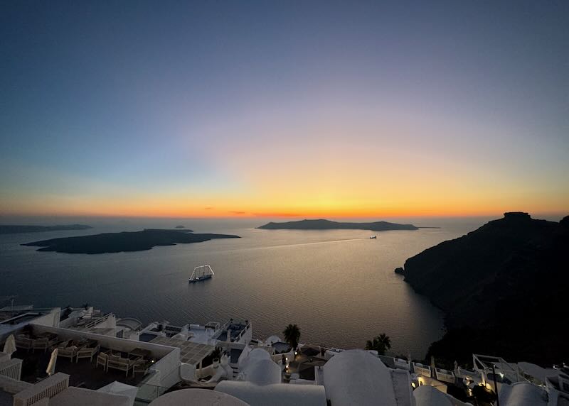 View of the Santorini caldera at sunset, with two ships leaving long wakes as they navigate away.