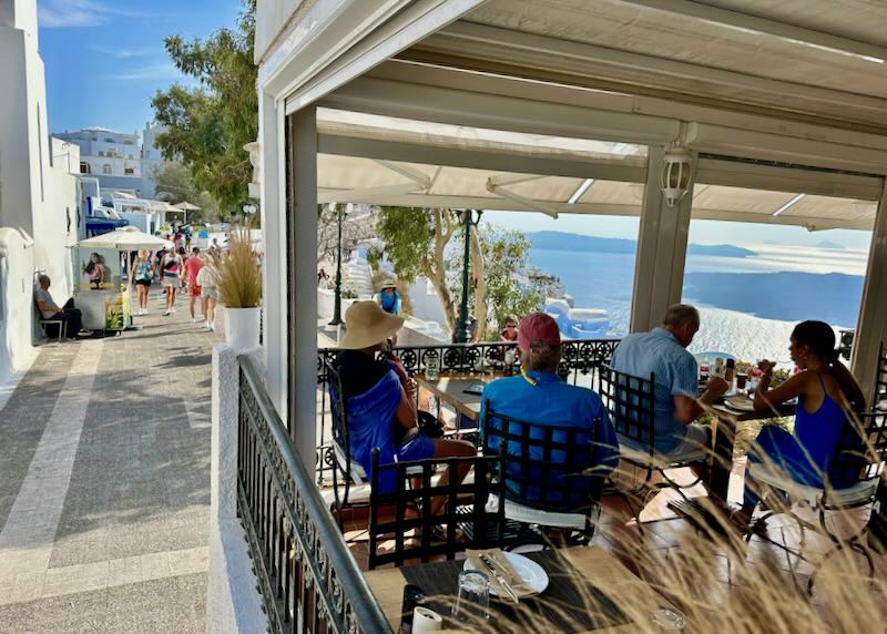 People sit at cafe tables perched near the edge of the caldera cliff in Firostefani, Santorini.