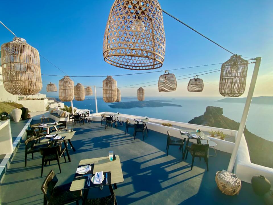 View over a dining terrace overhung by woven lanterns, overlooking the Santorini caldera.