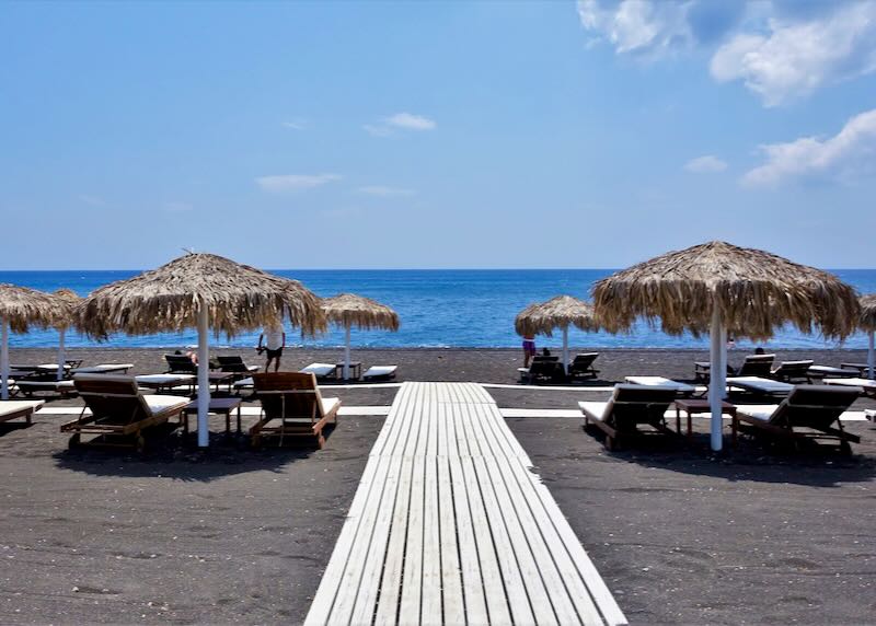 Beach track leading through black sand past thatched umbrellas to the sea.