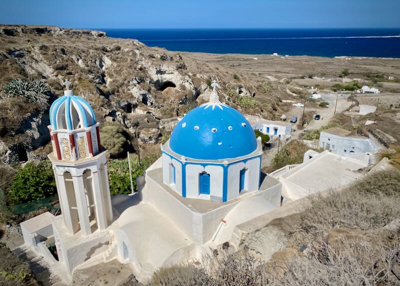 Bllue-domed church in the wilderness of Thirassia, covered in low-lying shrubs with cliffs, a the caldera spreading below, and Oia village in Santorini in the distance.