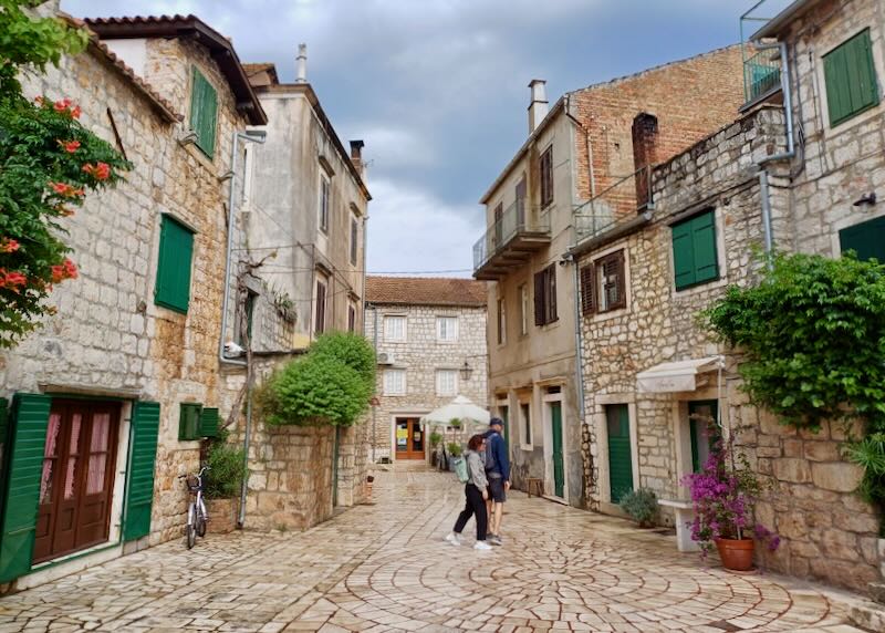 A couple wanders a cream-colored stone walkway with buildings the same color and stone with green shutters.