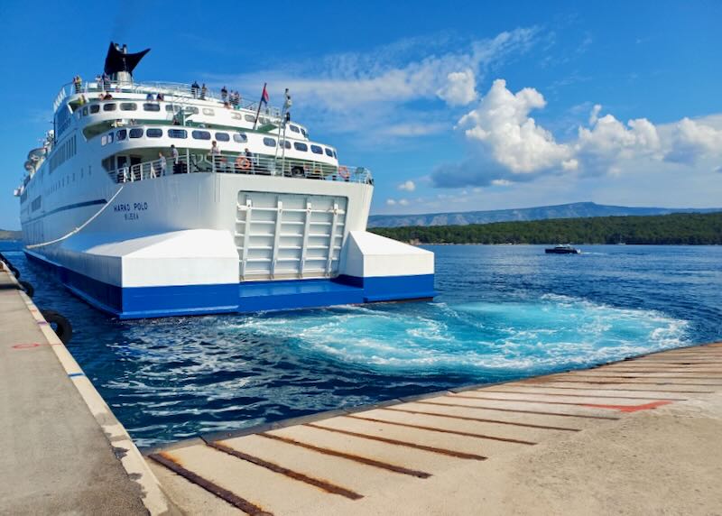 A large ferry produces light blue and white waves as it moves away from shore.