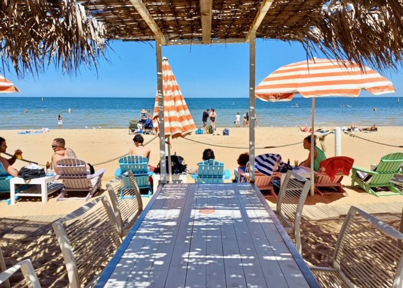 A long table and chairs sit on the beach under a grass thatched roof.