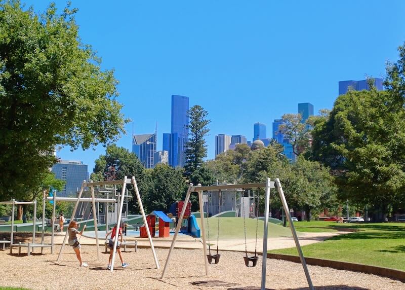 A couple of swing sets sit in a park with trees and views of the city.