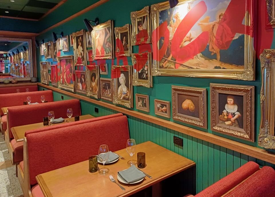 A wall of booths is lined with paintings in gold frames with red paint written over the top of them all.