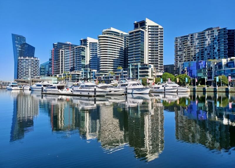 Tall city buildings reflect on the water by a harbor with boats.