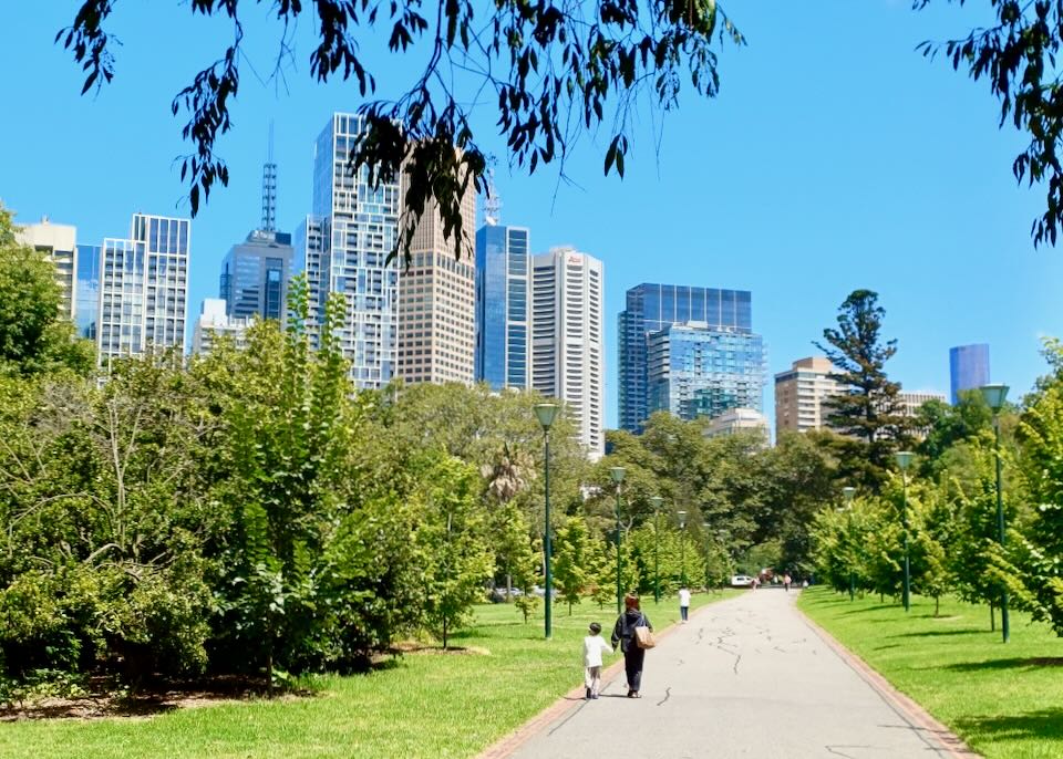 A few people walk through a park on a sunny day in a city with tall buildings.