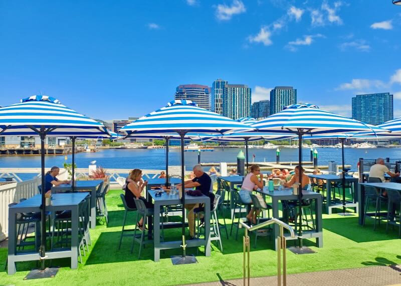 People sit under umbrellas at tables by a river.