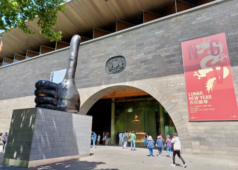 A large brass sculpture of a hand with an exaggerated thumb up sits outside a gallery.