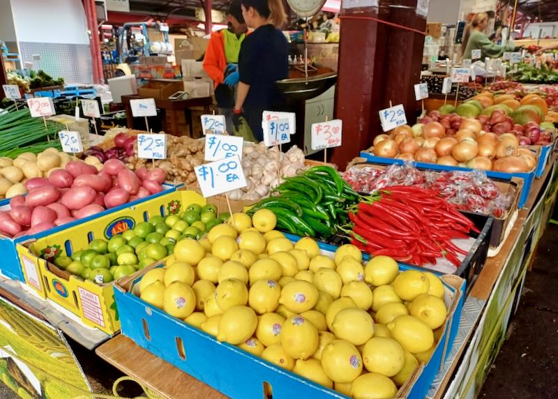 Bins of yellow lemons, green limes, red chili peppers, and potatoes, and onions sit at a market.