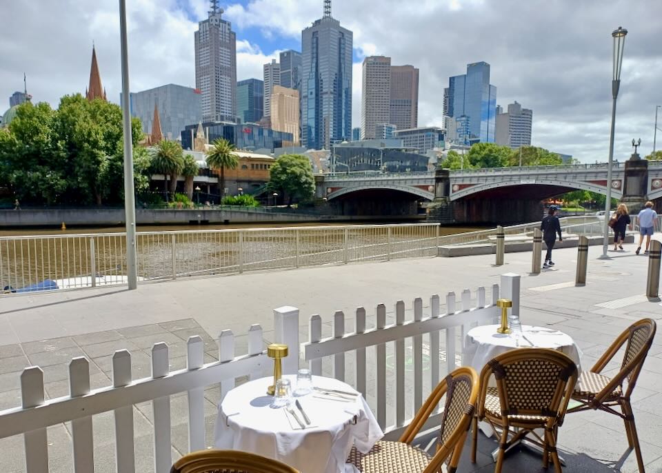 Dining tables sit by a river with a view of a city skyline.