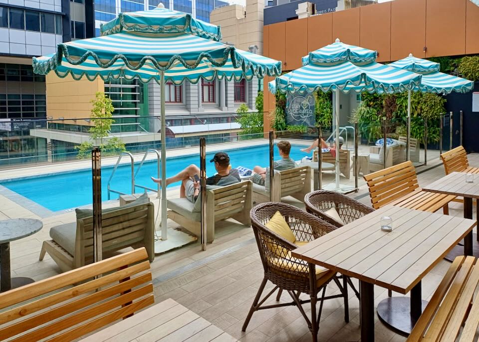 Three people sit by a small pool with blue and white stripped umbrellas and read their phones.