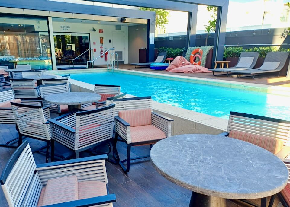 A long outdoor pool sits in the shade of a hotel with table and chairs next to it.