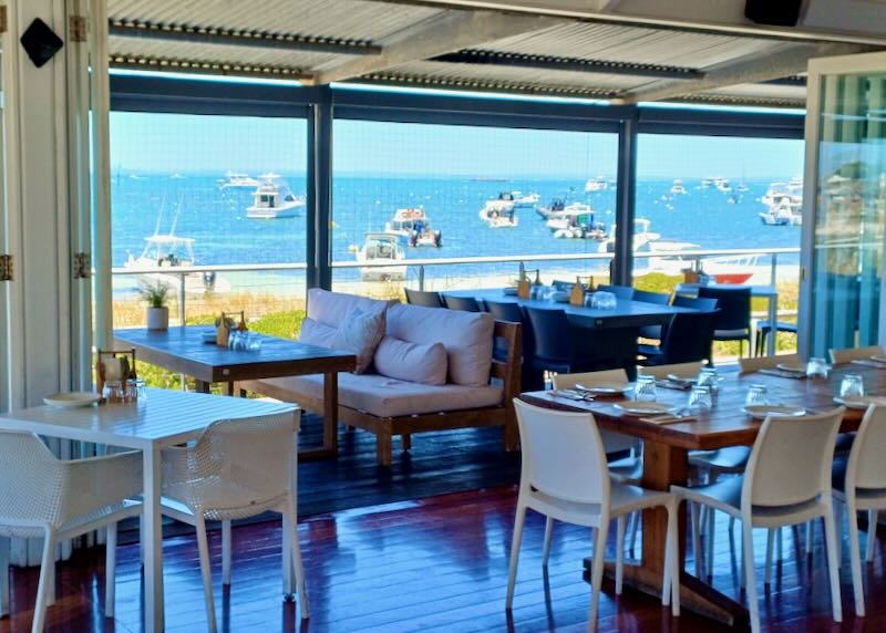 Table inside a restaurant overlook the ocean shore filled with boats.