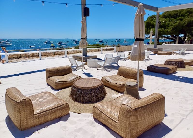 Large brown woven chairs sit on sand at a club next to the beach.