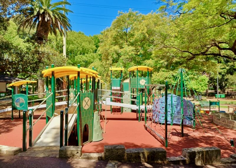 Lots of children's bright green playground equipment stands on rust colored sand.