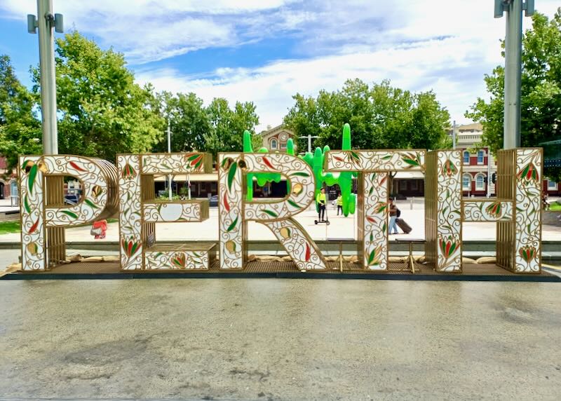 A large white sign with gold curls and gold edges sits by a park.