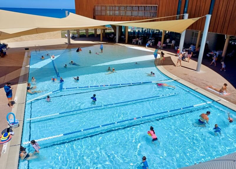 Children in colorful UV tops swim in a large pool.