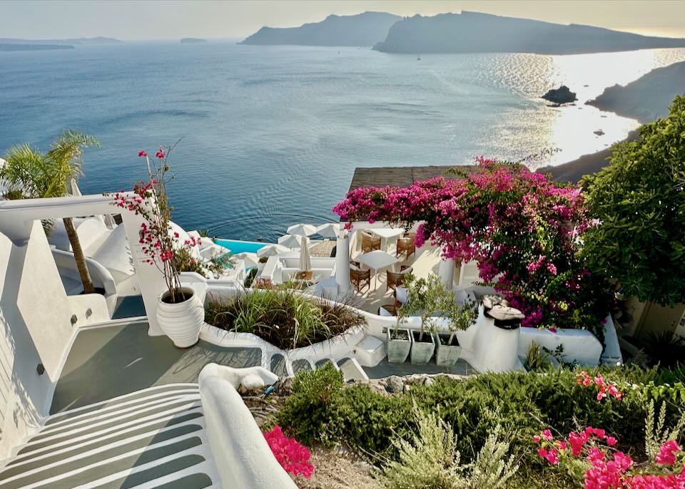 View down the white stairway of a cliffside hotel in Santorini, lined with bright flowers and overlooking the Santorini Caldera.