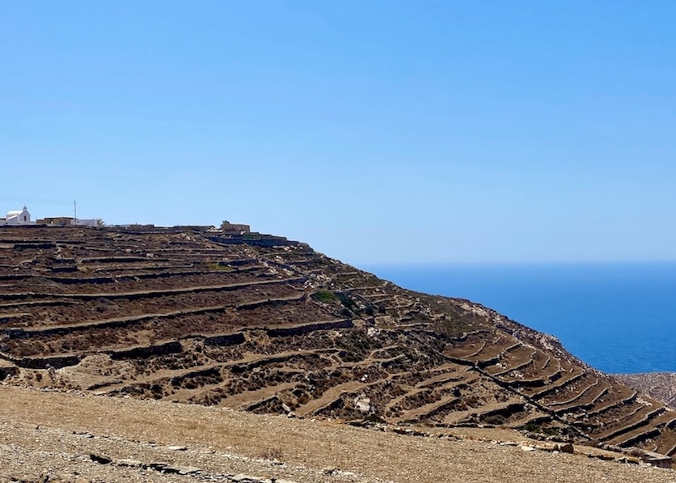 A farm spreads down the hill in rugged terraces with a white church on top and the sea in the distance in Ano Meria, Folegandros.