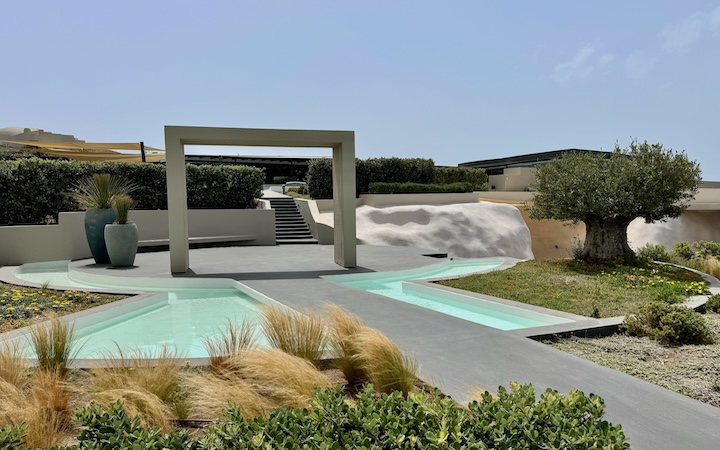 A freestanding, flat arch set on a dais with shallow pools on either side and an olive tree at the entrance of Divine Cave Experience in Imerovigli, Santorini.