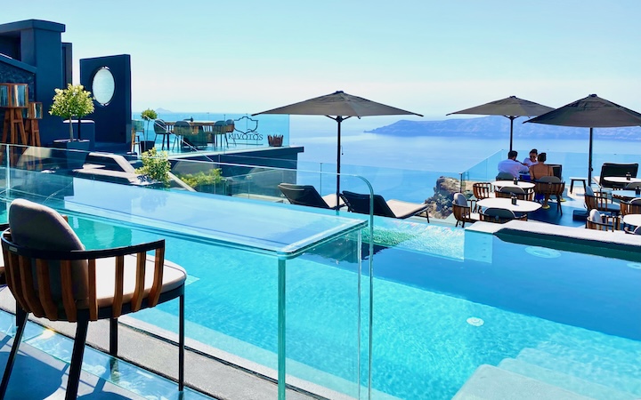 View from behind a glass table facing a glass-front infinity pool overlooking a the caldera at Kivotos Hotel in Imerovigli, Greece.