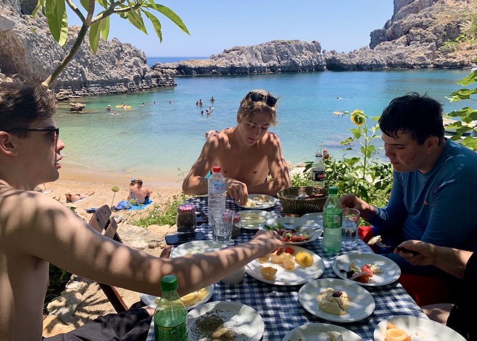 Teenagers having lunch at a picnic table on the beach in front of a blue sea at St. Paul's Bay in Lindos, Rhodes.