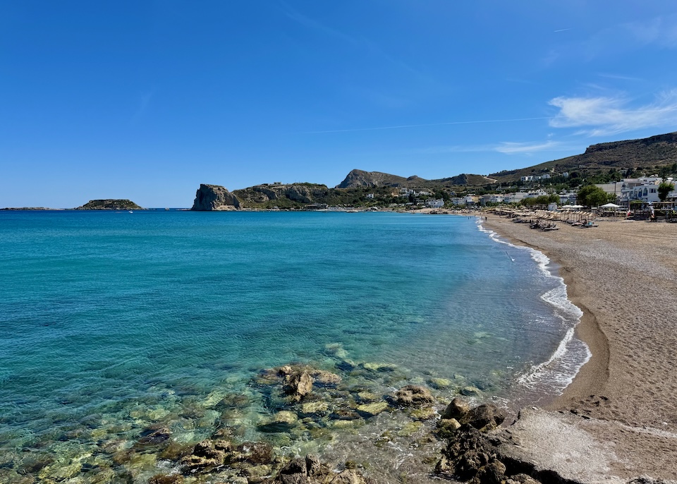 A long, sandy beach with stones in the foreground and rows of sunbeds and umbrellas in the distance at Segna Beach in Rhodes.